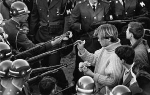 a protestor places a flower in the rifle barrel of a police officer